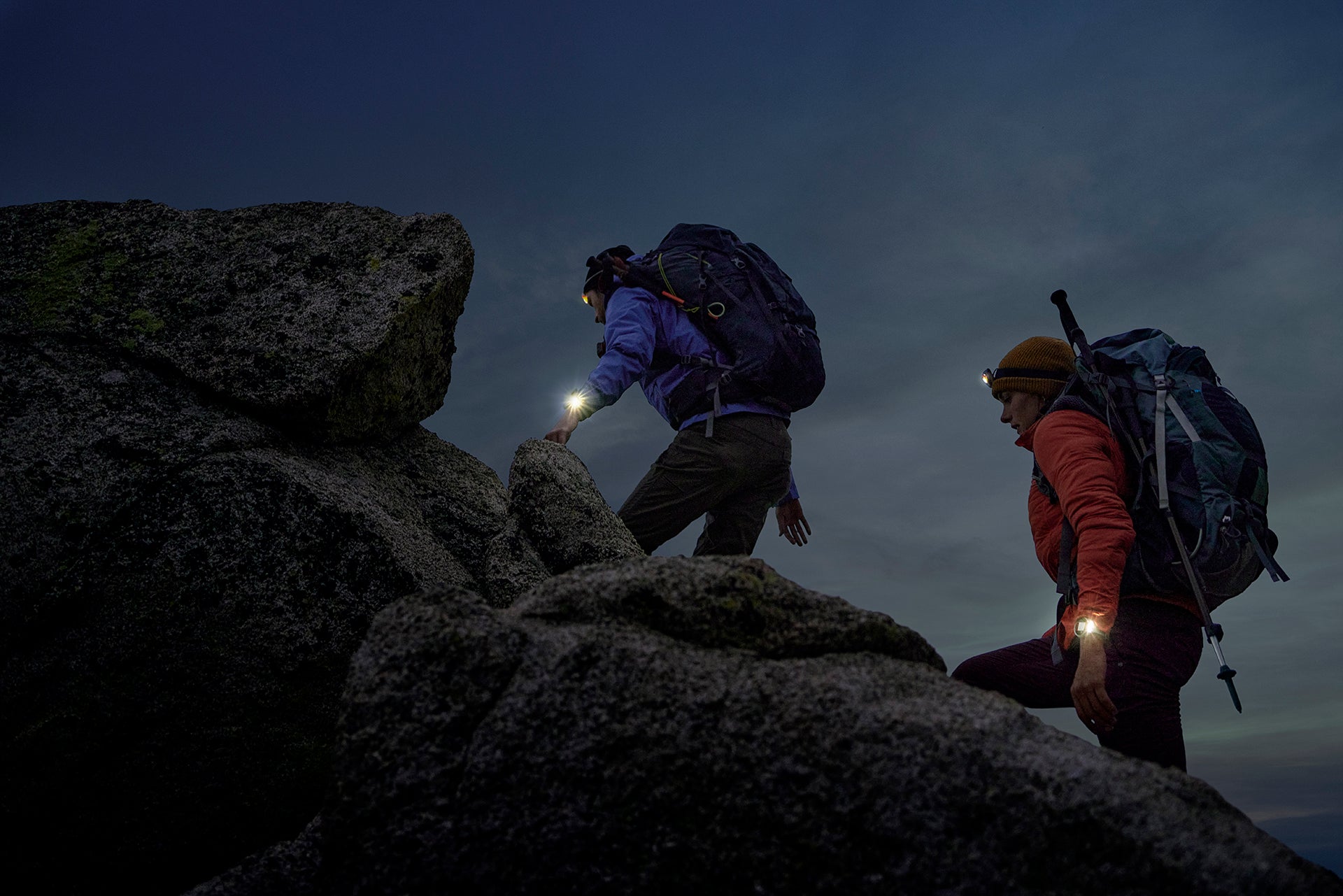 Two hikers going up a rocky trail at dusk with wearing backpacks, headlamps and fenix 7 watches with the LED flashlights on