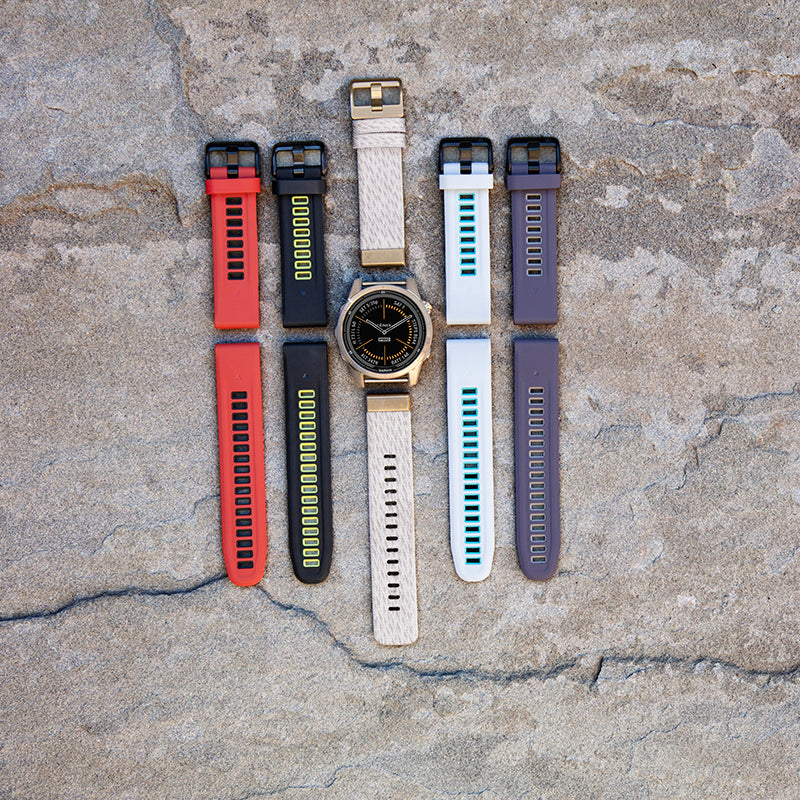 The Garmin fenix 7S soft gold/light sand GPS watch with four other watch bands (red, black, white, purple) all laying flat on a rock